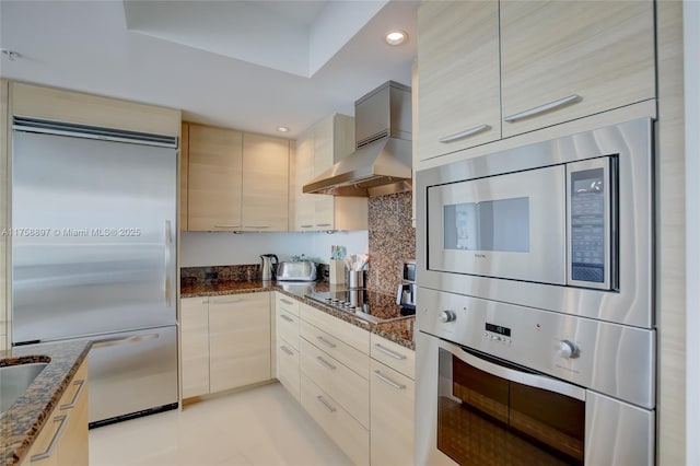 kitchen featuring dark stone counters, built in appliances, modern cabinets, and wall chimney range hood