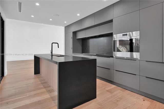 kitchen with light wood-style flooring, an island with sink, a sink, oven, and modern cabinets