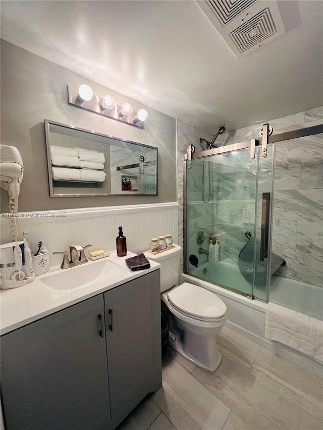 full bath featuring visible vents, bath / shower combo with glass door, toilet, wainscoting, and vanity