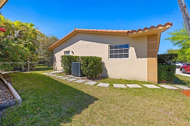 view of side of property featuring fence, stucco siding, central air condition unit, a tiled roof, and a lawn