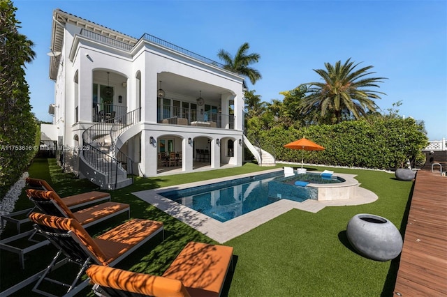 rear view of property with stairway, a balcony, an in ground hot tub, a patio area, and a lawn