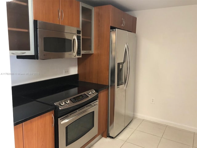 kitchen featuring brown cabinets, dark countertops, appliances with stainless steel finishes, light tile patterned floors, and glass insert cabinets