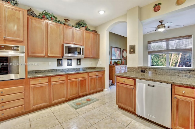 kitchen with a ceiling fan, light stone counters, stainless steel appliances, arched walkways, and light tile patterned flooring