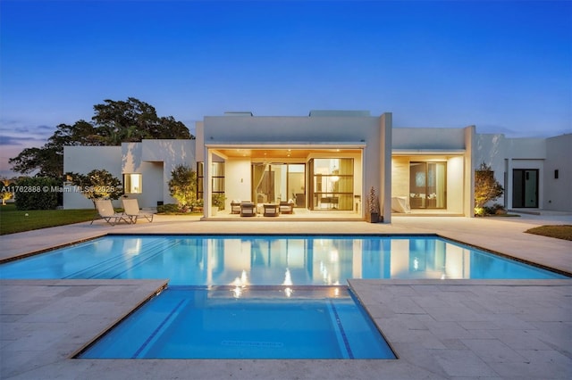 back of property at dusk featuring stucco siding, a pool with connected hot tub, and a patio