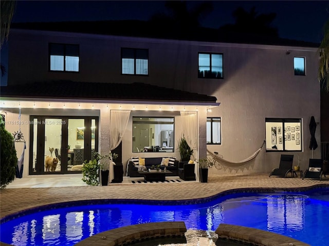 back of house at night featuring an outdoor living space, a patio area, a pool with connected hot tub, and stucco siding