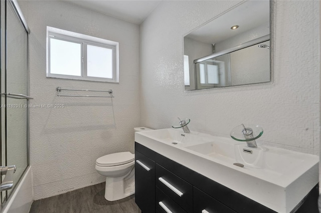 bathroom featuring toilet, wood finished floors, double vanity, a textured wall, and combined bath / shower with glass door