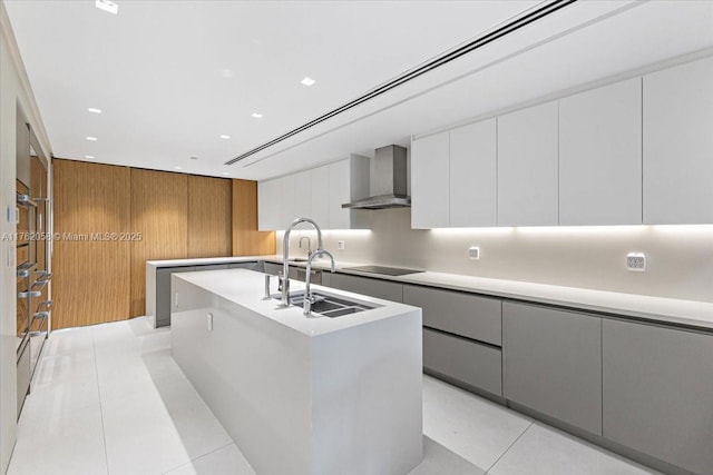 kitchen featuring modern cabinets, an island with sink, wall chimney exhaust hood, light countertops, and black electric cooktop