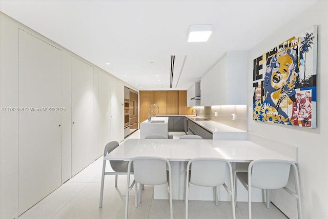 kitchen featuring modern cabinets, a peninsula, a breakfast bar area, wall chimney range hood, and light countertops