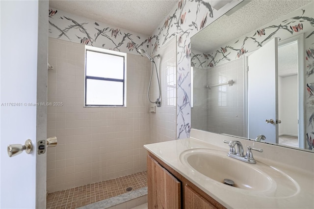 bathroom featuring wallpapered walls, vanity, tiled shower, and a textured ceiling