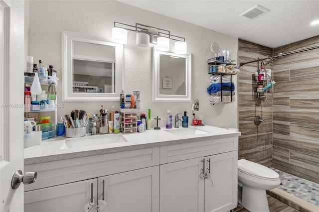 bathroom with visible vents, toilet, a sink, a tile shower, and double vanity