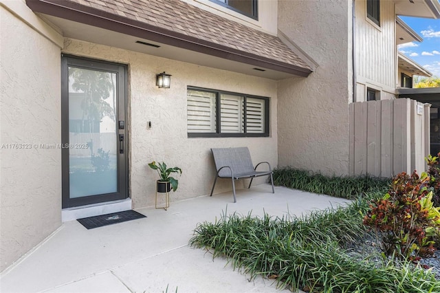 entrance to property with a shingled roof, a patio, fence, and stucco siding