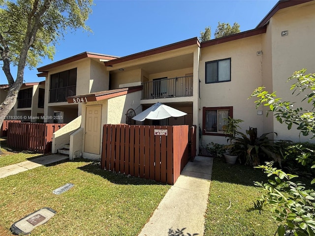 view of front of house featuring stucco siding, a front lawn, a balcony, and fence