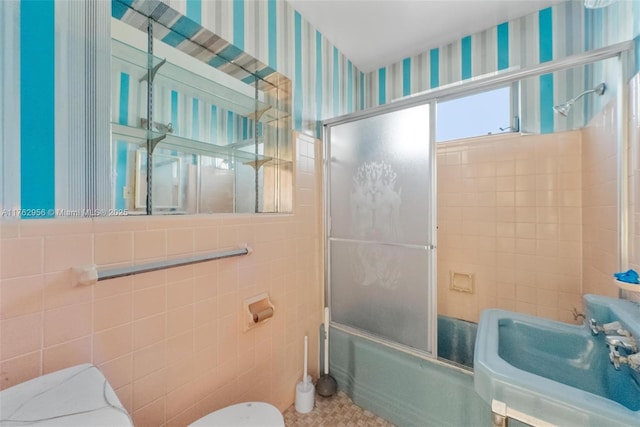 bathroom featuring wallpapered walls, toilet, shower / bath combination with glass door, tile walls, and a sink