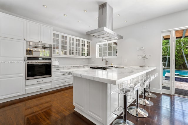 kitchen with glass insert cabinets, dark wood-type flooring, light stone countertops, a kitchen breakfast bar, and island exhaust hood