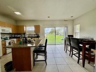 kitchen featuring white appliances, light tile patterned floors, brown cabinetry, dark countertops, and a center island