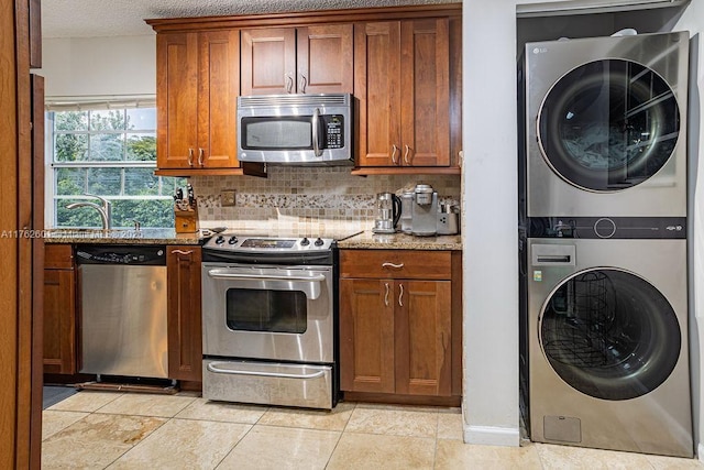 kitchen with stacked washer and dryer, a sink, appliances with stainless steel finishes, brown cabinetry, and decorative backsplash