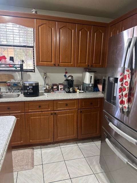 kitchen with a sink, stainless steel fridge, brown cabinetry, light tile patterned floors, and light stone countertops