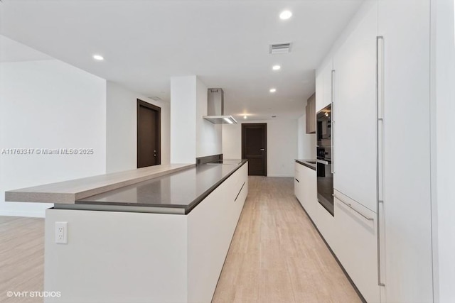 kitchen with dark countertops, light wood-type flooring, white cabinets, modern cabinets, and wall chimney exhaust hood