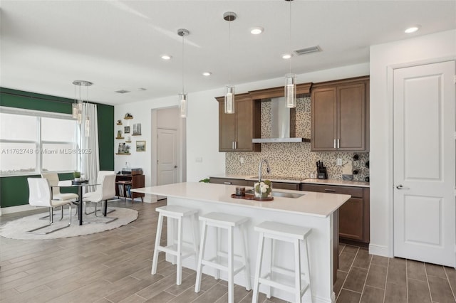 kitchen with visible vents, an island with sink, backsplash, wall chimney exhaust hood, and light countertops