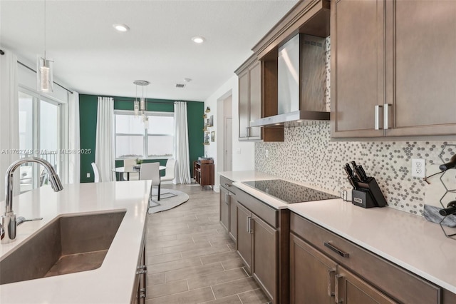 kitchen with a sink, tasteful backsplash, wall chimney range hood, black electric stovetop, and hanging light fixtures