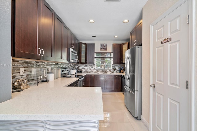 kitchen featuring light stone countertops, a peninsula, decorative backsplash, dark brown cabinets, and appliances with stainless steel finishes