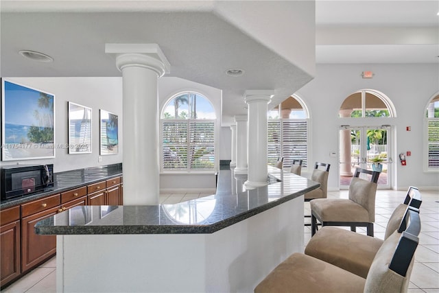 kitchen with brown cabinetry, ornate columns, light tile patterned flooring, black microwave, and open floor plan