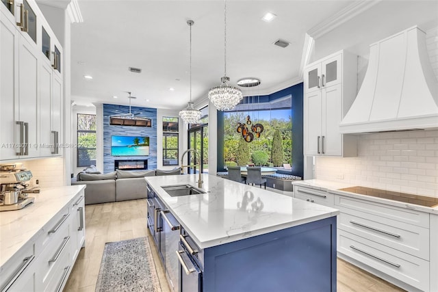 kitchen with black electric stovetop, premium range hood, a center island with sink, ornamental molding, and a sink