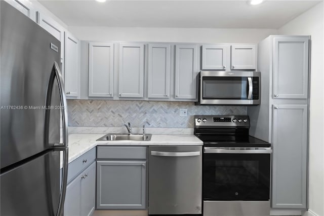 kitchen with gray cabinetry, a sink, backsplash, appliances with stainless steel finishes, and light stone countertops