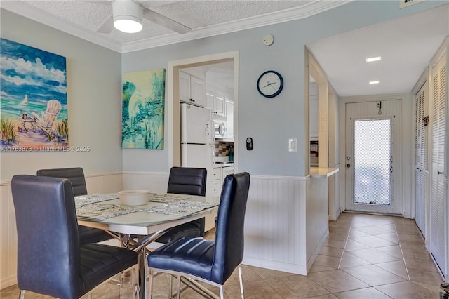 dining space with a ceiling fan, a textured ceiling, wainscoting, crown molding, and light tile patterned floors