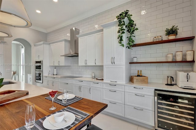 kitchen with stainless steel double oven, arched walkways, light countertops, wine cooler, and wall chimney range hood