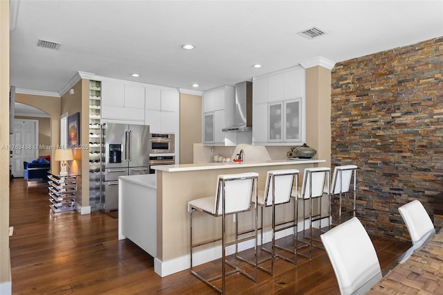 kitchen featuring a peninsula, wall chimney range hood, visible vents, and appliances with stainless steel finishes