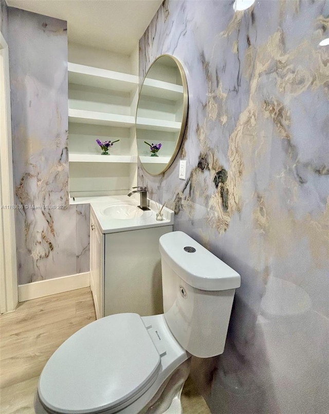 half bath featuring wallpapered walls, baseboards, toilet, wood finished floors, and vanity