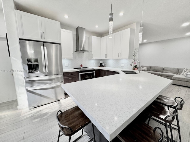 kitchen with a breakfast bar area, a peninsula, a sink, stainless steel appliances, and wall chimney exhaust hood