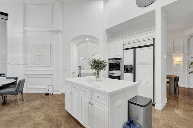 kitchen featuring stainless steel double oven, arched walkways, white cabinetry, a decorative wall, and a center island