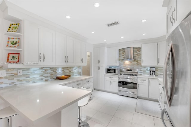 kitchen featuring visible vents, a kitchen breakfast bar, a peninsula, appliances with stainless steel finishes, and wall chimney exhaust hood