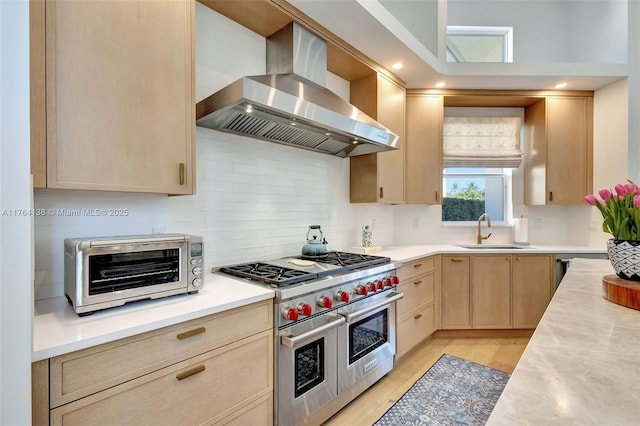 kitchen with light brown cabinets, a sink, wall chimney range hood, light countertops, and range with two ovens