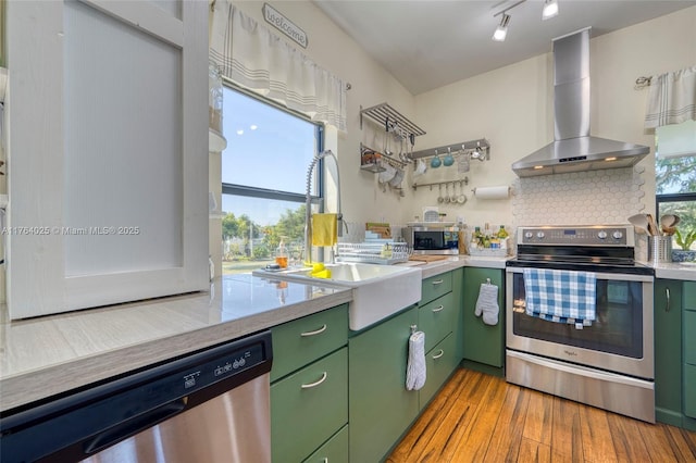 kitchen with light wood-style flooring, stainless steel appliances, green cabinets, wall chimney range hood, and tasteful backsplash