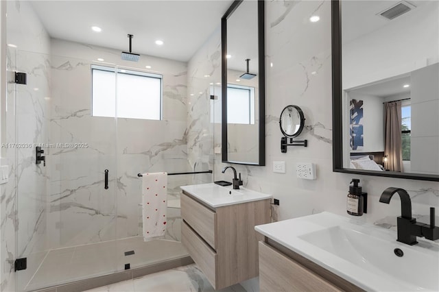 full bathroom with visible vents, two vanities, a sink, a marble finish shower, and tile walls