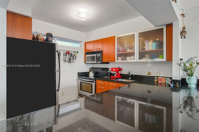 kitchen with glass insert cabinets, dark stone counters, brown cabinets, appliances with stainless steel finishes, and a sink