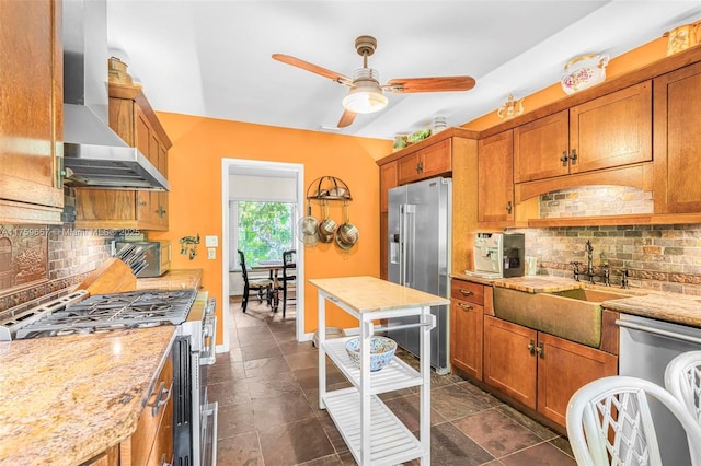 kitchen featuring wall chimney range hood, brown cabinetry, stainless steel appliances, a ceiling fan, and a sink