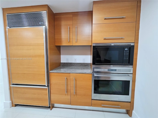 kitchen with brown cabinets, dark countertops, built in appliances, light tile patterned floors, and baseboards