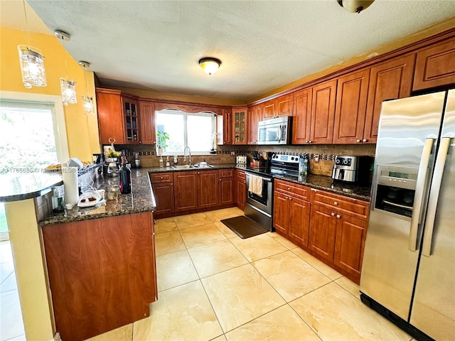 kitchen with a sink, appliances with stainless steel finishes, glass insert cabinets, brown cabinetry, and dark stone counters