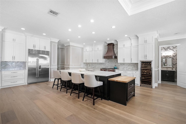 kitchen featuring light wood-style floors, beverage cooler, premium range hood, a large island with sink, and built in refrigerator