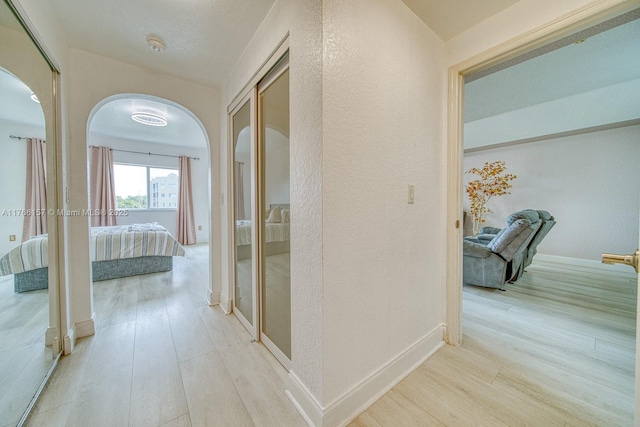 hallway with arched walkways, a textured wall, baseboards, and light wood-type flooring