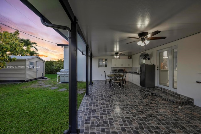 patio terrace at dusk with an outbuilding, a lawn, area for grilling, a shed, and ceiling fan