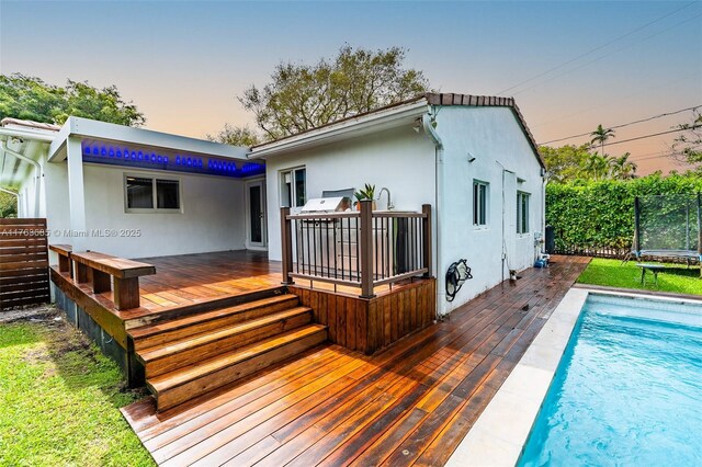 back of house at dusk with stucco siding, a trampoline, a deck, and an outdoor pool