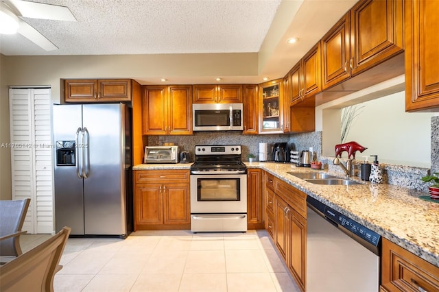 kitchen with a sink, light stone counters, stainless steel appliances, brown cabinetry, and light tile patterned floors