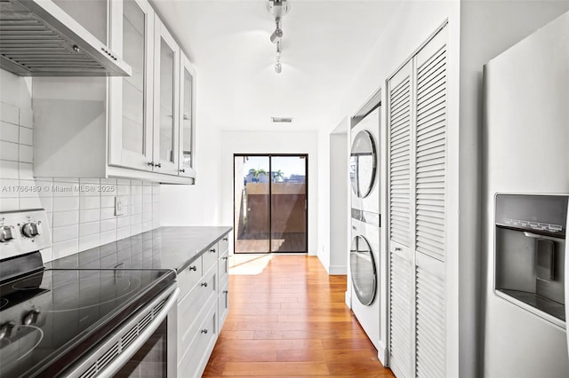 kitchen with light wood-style flooring, stainless steel electric range, stacked washer and dryer, dark countertops, and wall chimney exhaust hood