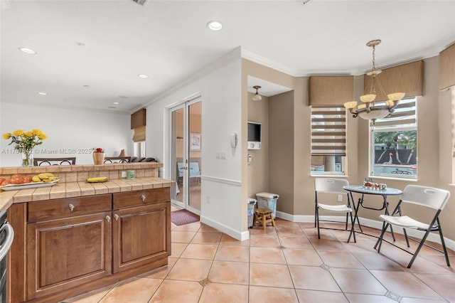 kitchen with light tile patterned floors, an inviting chandelier, tile counters, crown molding, and brown cabinets