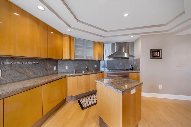kitchen featuring light wood finished floors, wall chimney range hood, dark stone countertops, a raised ceiling, and a sink